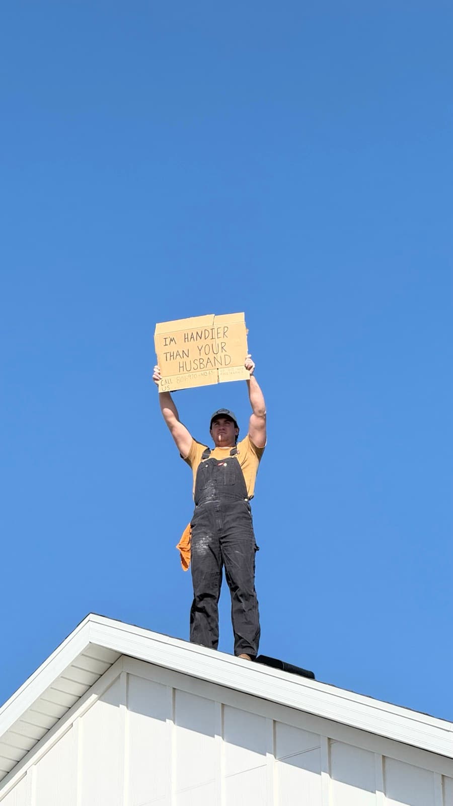 Cory, founder of Planned Home Maintenance, standing on a rooftop holding a sign reading 'I'm handier than your husband'